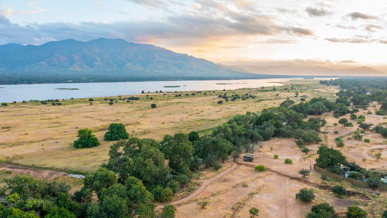 Little Mana Sands Aerial - A golden-hour aerial view of Little Mana Sands, nestled discreetly beneath the tree line with sweeping views of the Zambezi River and escarpment. The camp’s rustic charm blends seamlessly into the floodplain, offering guests an intimate and immersive safari setting.